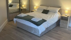 Ein ordentlich bezogenes Bett mit grüner Tagesdecke, zwei Nachttische mit Lampen, ein großer Spiegel und ein Holzschrank.
