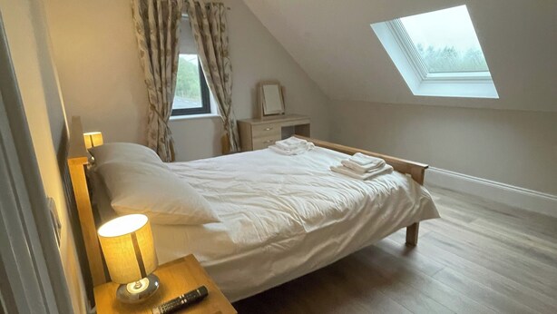 Ein Schlafzimmer mit schrägen Wänden, einem Oberlicht, einem Bett mit weißer Bettwäsche, einem Nachttisch mit Lampe sowie einem Beistelltisch mit Fernbedienung.