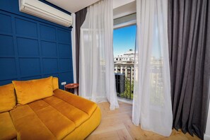 A room with a yellow sofa, a small table, and a view of a cityscape through sheer curtains. - Blue Moon - One Bedroom - Romana Square (Bucharest)
