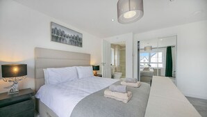 A modern hotel room with a large bed, a bedside table with a lamp, a painting on the wall, and a view of the bathroom through an open door. - Fern Place Villa  | Grampian Lettings Ltd (Aberdeen City)