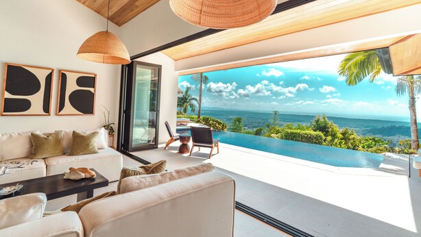 A modern living room with a large window overlooking a private pool and ocean view. - Arbórea Villas (Ciudad Cortés)