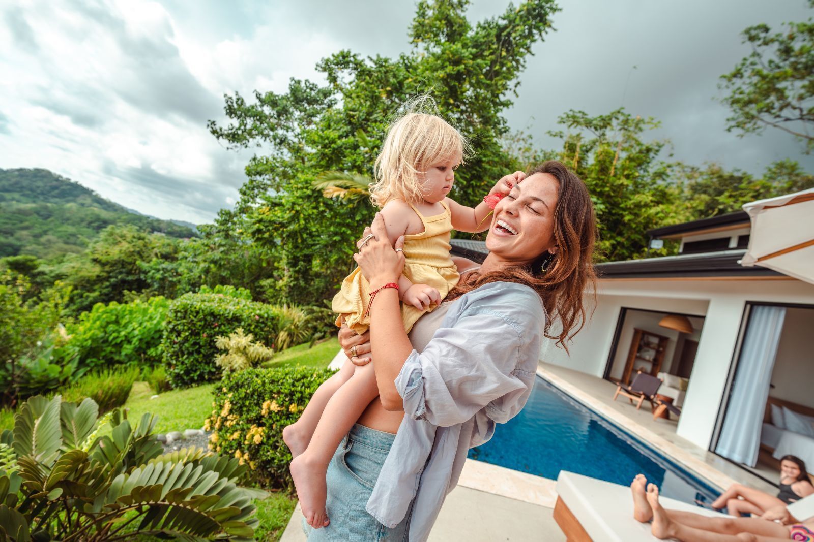 A woman holding a child by a poolside with a view of lush greenery and a modern building in the background.