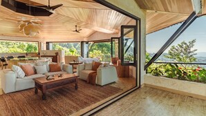 A spacious living area with a wooden ceiling, a large window offering a view of greenery, and a kitchen area with a bar. - Arbórea Villas (Ciudad Cortés)