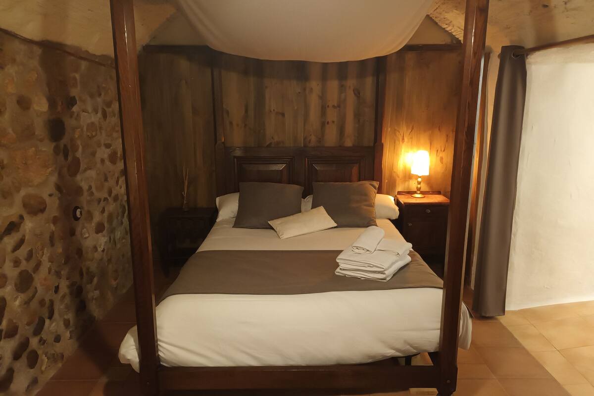 A four-poster bed with a canopy, a bedside table with a lamp, and a stone wall.