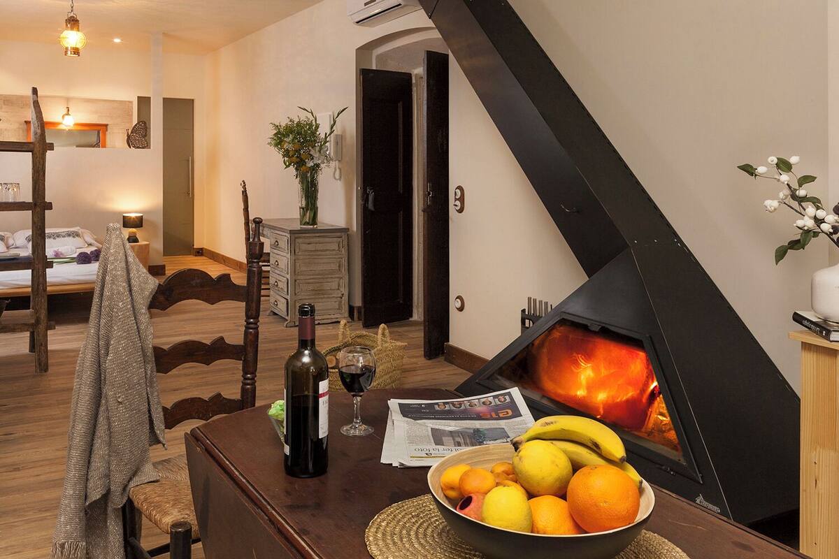 A cozy living room with a fireplace, a dining table with fruits and wine, and a bed in the background.