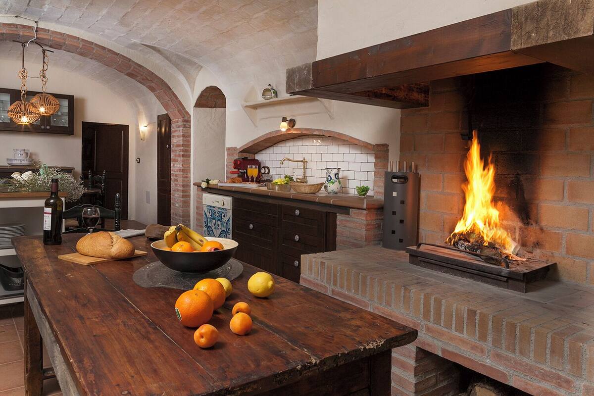 A rustic kitchen with a brick fireplace, wooden table, and a bowl of fruit.