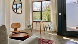 Une pièce moderne avec une table ronde en bois, deux chaises en métal, un canapé gris, un tapis à motifs rouges, une grande fenêtre avec des rideaux et une porte dotée d’une poignée.