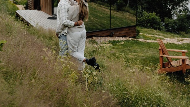 Un couple s’embrasse devant une structure moderne en bois dotée d’une grande surface réfléchissante, surplombant une étendue d’eau et entourée de verdure.