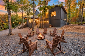 A cabin with a fire pit and outdoor seating, surrounded by trees and a gravel ground.