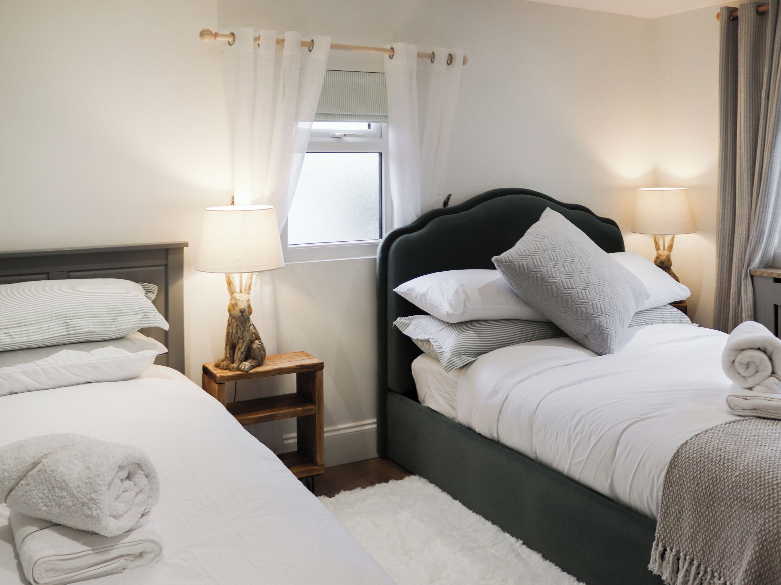 Una cama bien hecha con almohadas y una toalla enrollada, una mesita de noche con una lámpara y una ventana con cortinas.
