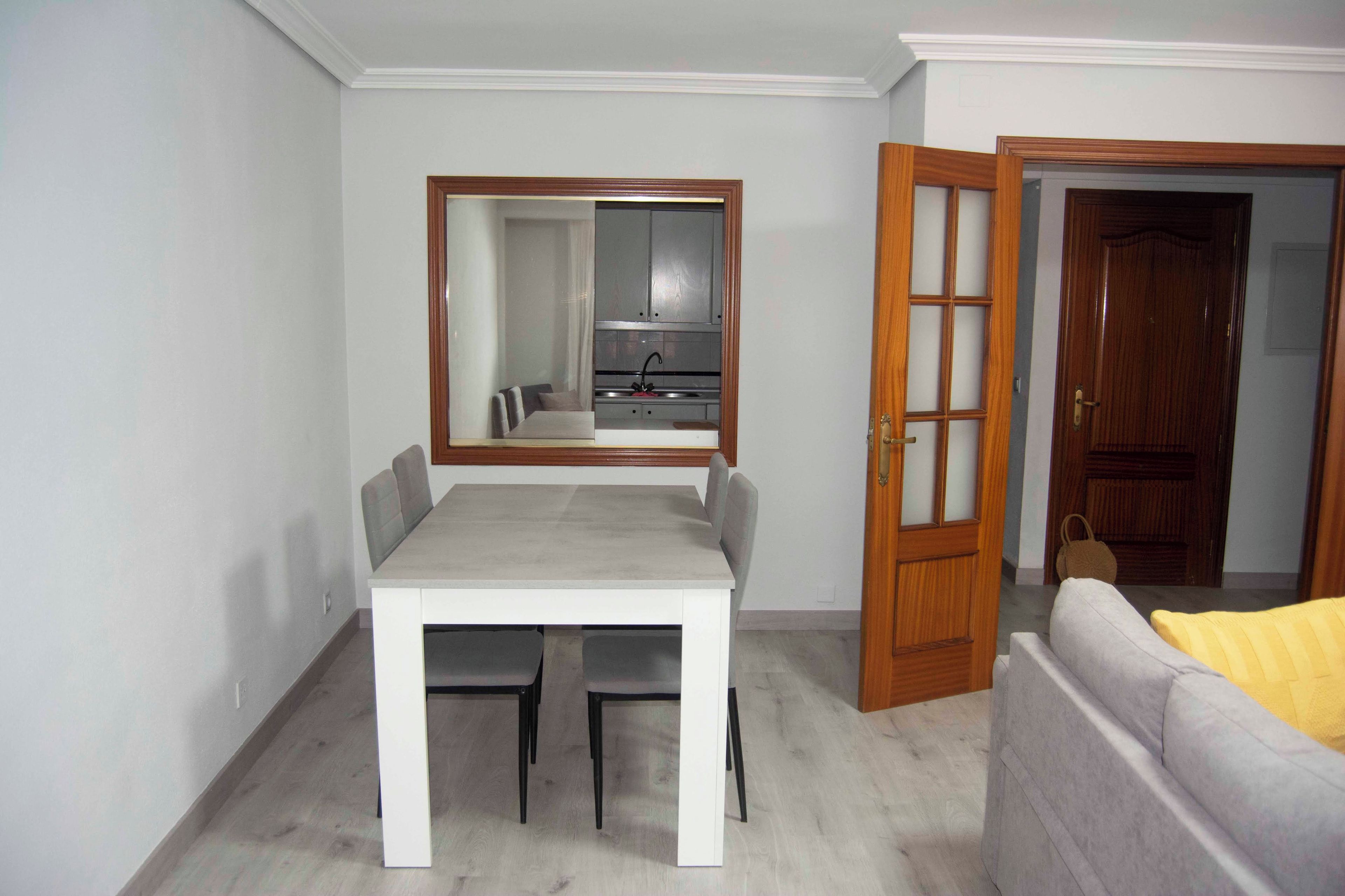 Una habitación pequeña con una mesa de comedor, dos sillas, un sofá, una puerta de madera y un espejo en la pared.