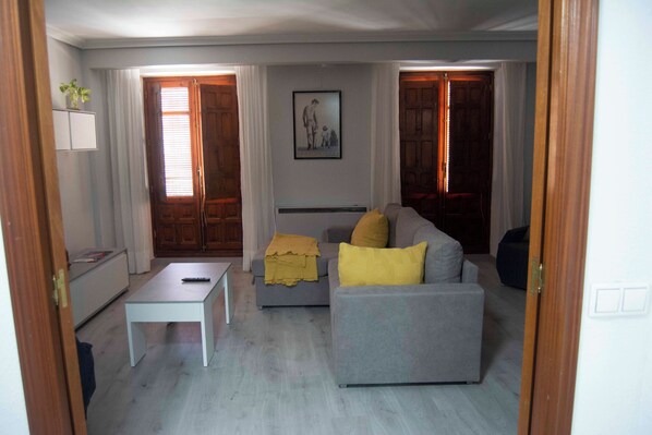 A living room with a grey sofa, a small white table, and a yellow throw blanket. There is a framed picture on the wall and a window with curtains.