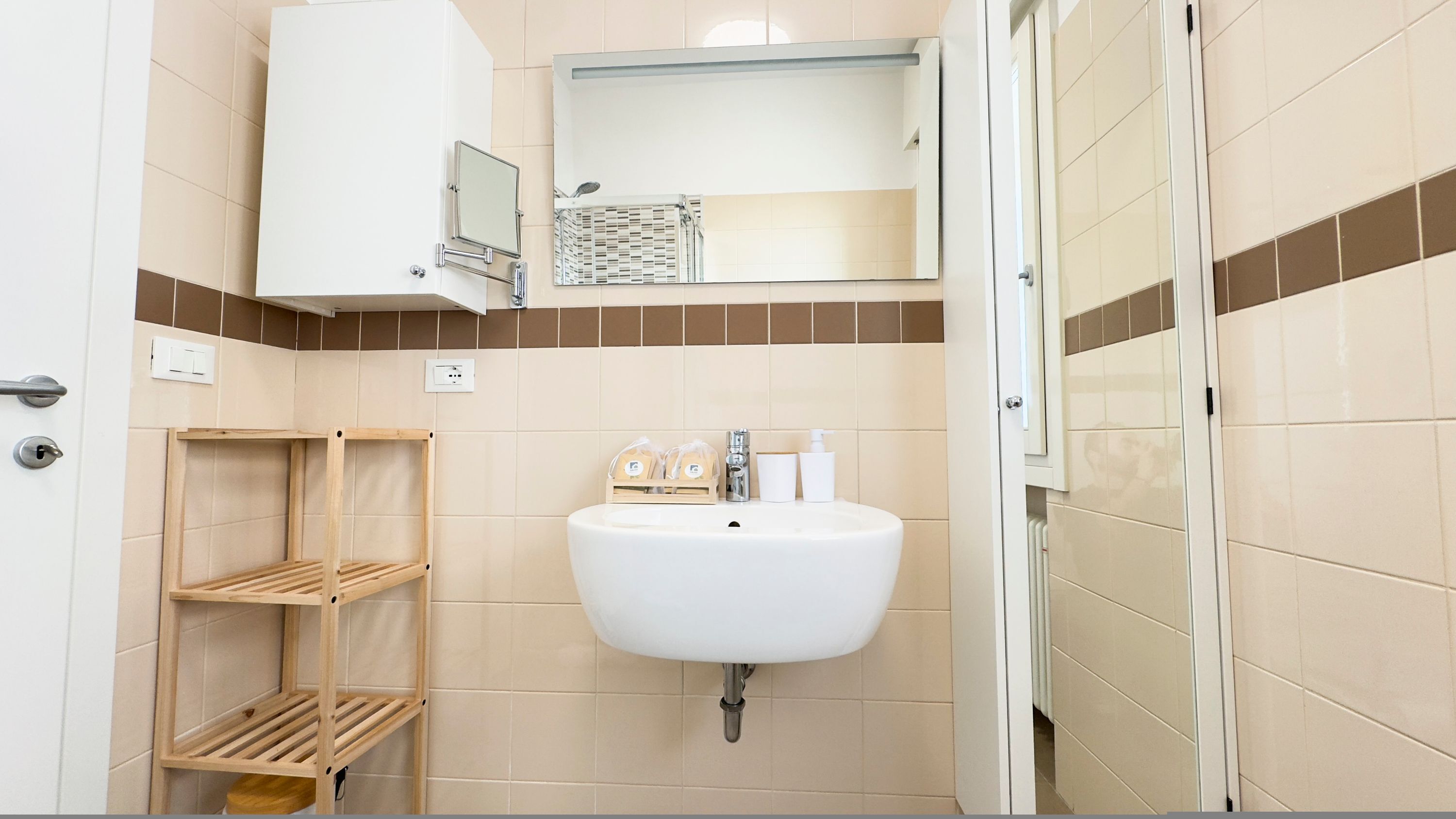Un baño con un lavabo blanco, un espejo encima y una zona de ducha. Hay estantes de madera y artículos de aseo en el lavabo.