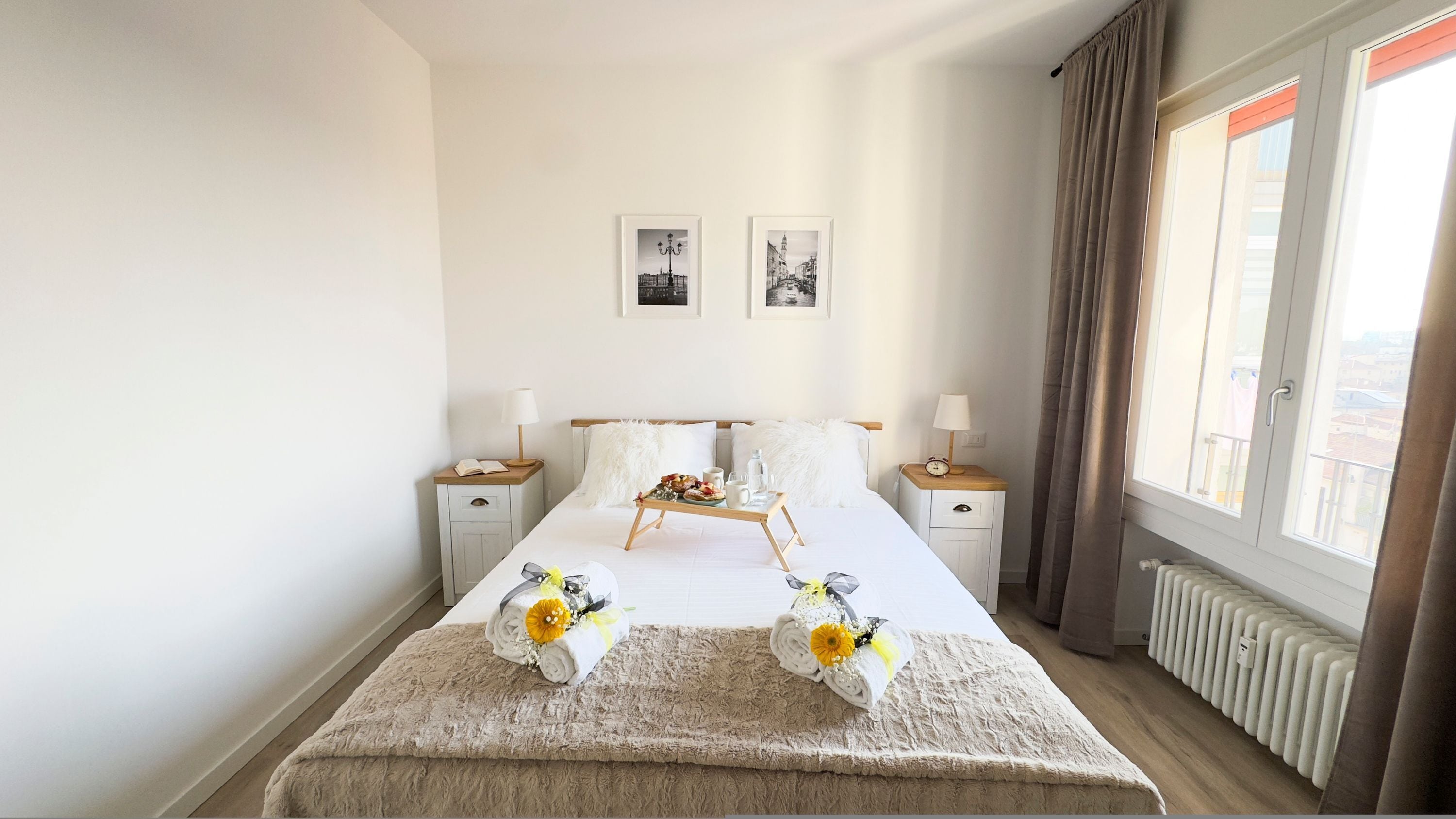 A neatly made bed with a fluffy beige blanket, two bedside tables with lamps, a tray with breakfast items, and two framed pictures on the wall.