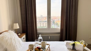 A hotel room with a bed, a bedside table with a lamp and alarm clock, a window with curtains, a radiator, and a table with breakfast items and a bouquet of flowers. - Venezia Dream Stay by Ca-sa Check (Mestre)