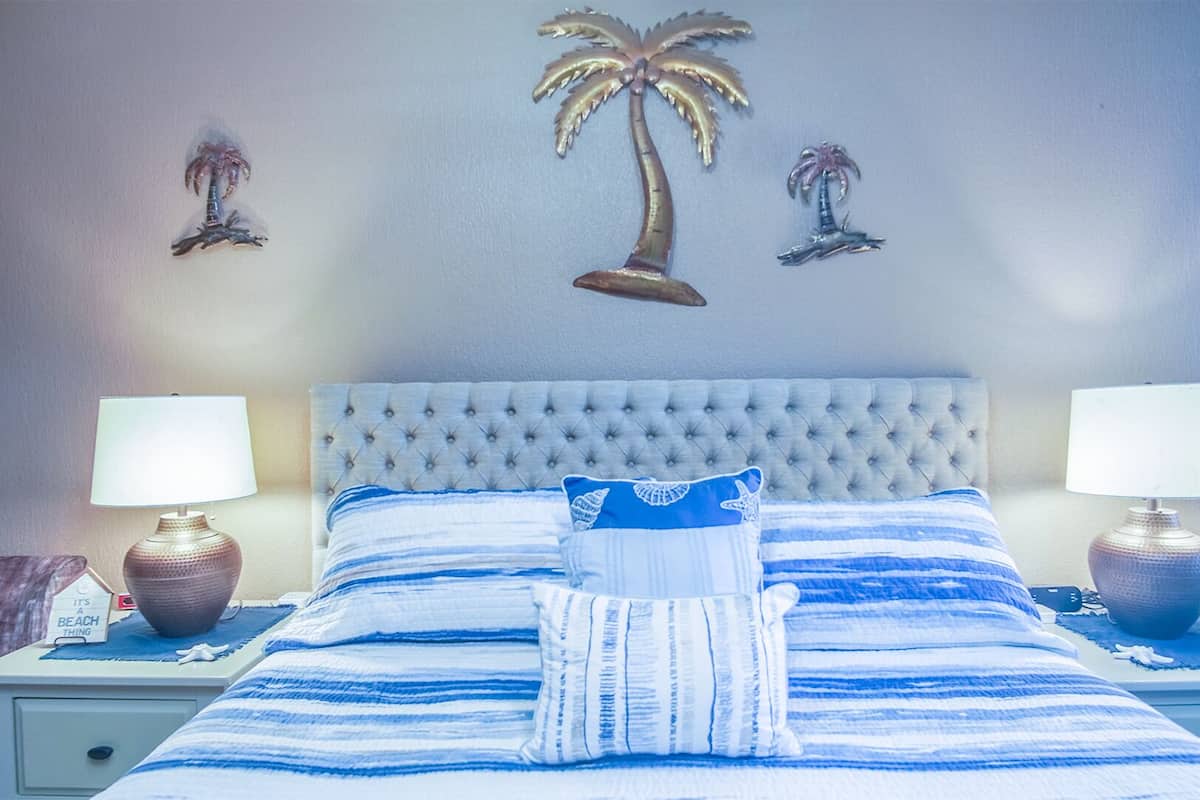 Une chambre d’hôtel avec un grand lit, deux lampes de chevet, une table de chevet et un mur décoré de trois motifs représentant des palmiers.