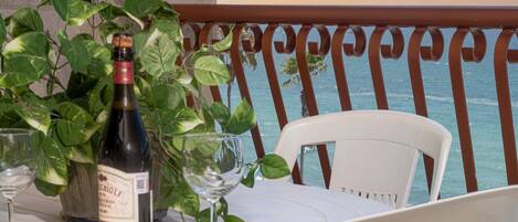 Una mesa con una botella de vino, dos copas de vino y una planta en maceta en un balcón con vistas al mar.