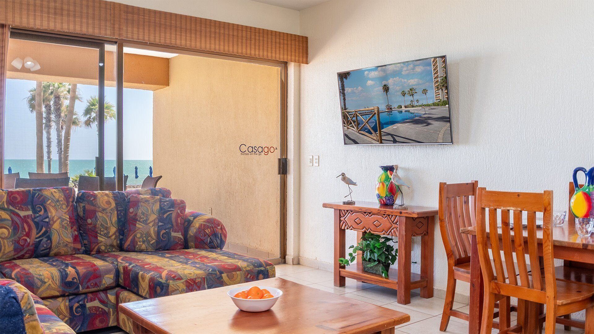 A living room with a colorful sofa, a wooden table with a bowl of fruit, a wooden chair, a small table with a vase, and a painting of a beach scene.
