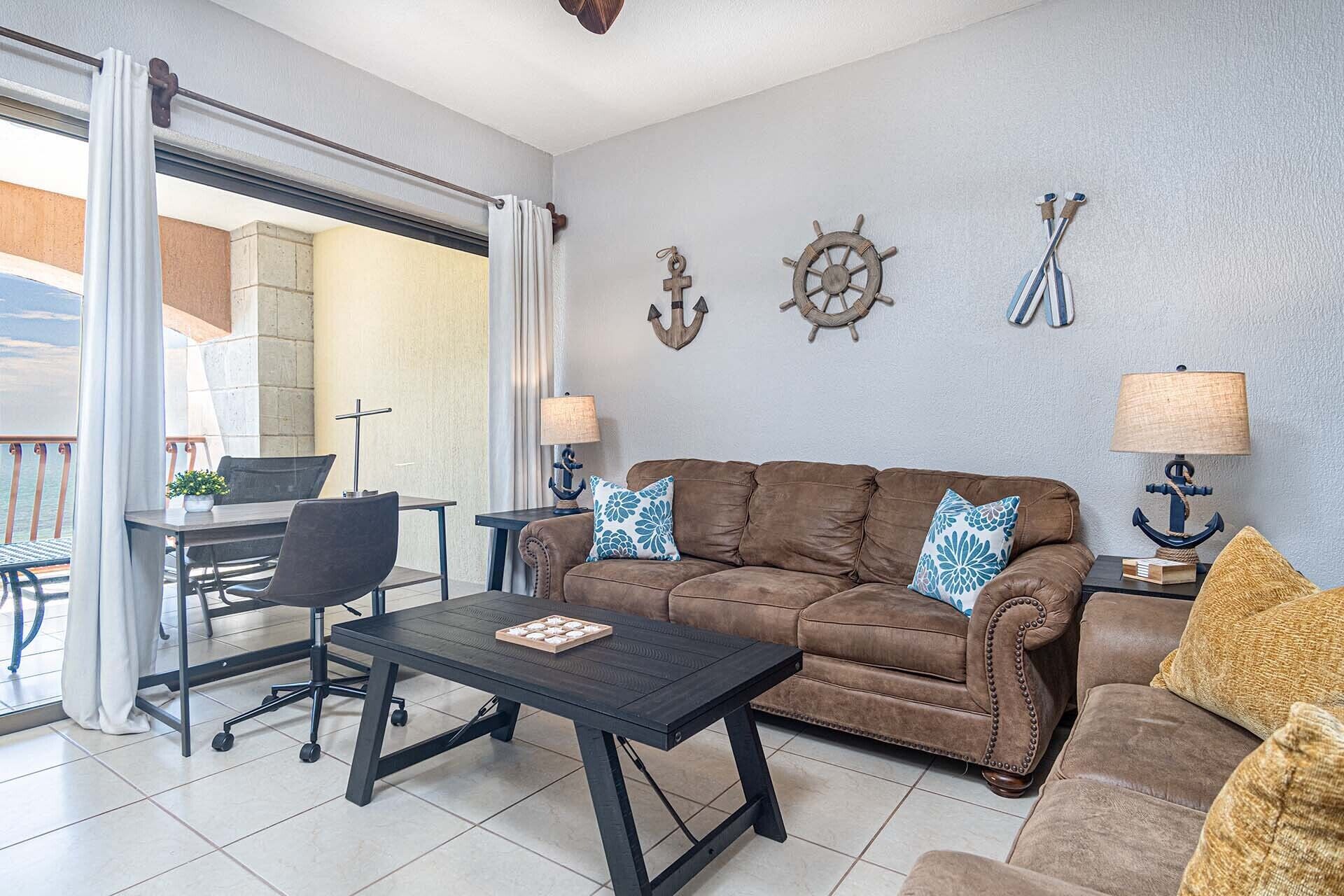 Un salon avec un canapé marron, une table basse, un coin repas avec une table et des chaises, et des éléments de décoration sur le thème de la marine sur les murs.