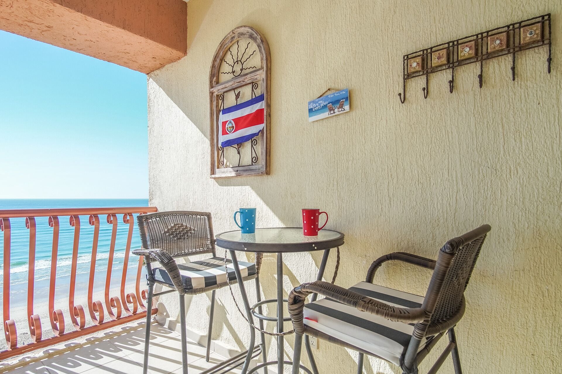 A balcony with wicker chairs, a small table, a flag, and a metal railing overlooking the sea.