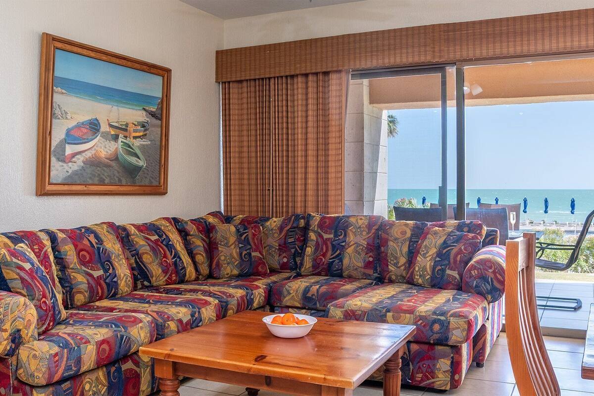 A beach-themed living room with a colorful sofa, a painting of boats on the beach, and a view of the ocean through large windows.