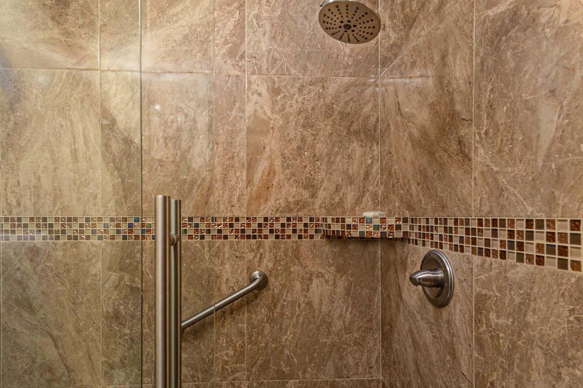 A shower area with a glass door, a handrail, a showerhead, and a tiled backsplash.