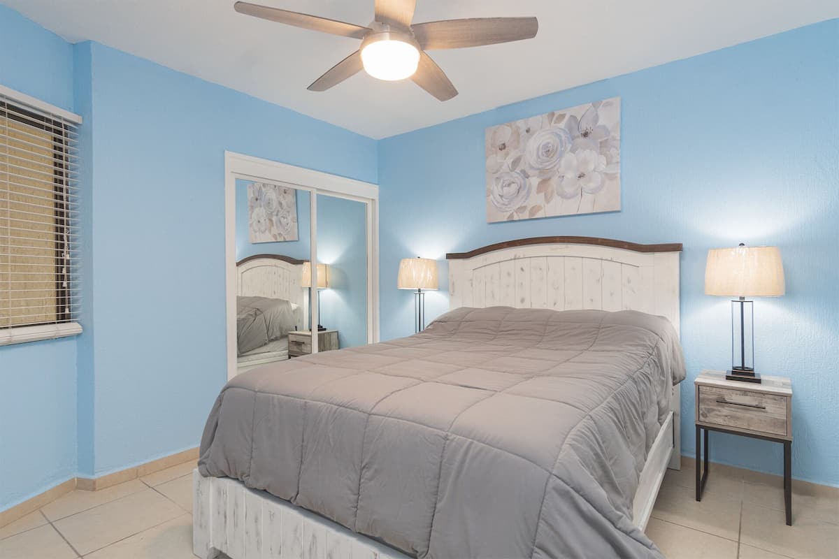 Une chambre à coucher avec un lit, deux tables de chevet avec des lampes, un ventilateur de plafond, une fenêtre avec des stores et une œuvre d’art florale accrochée au mur.