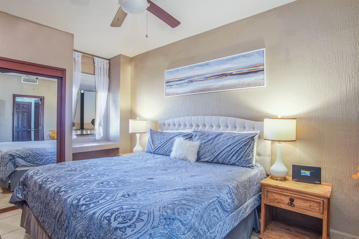 Une chambre d’hôtel avec un grand lit, deux tables de chevet avec des lampes, un ventilateur de plafond et un tableau au mur.