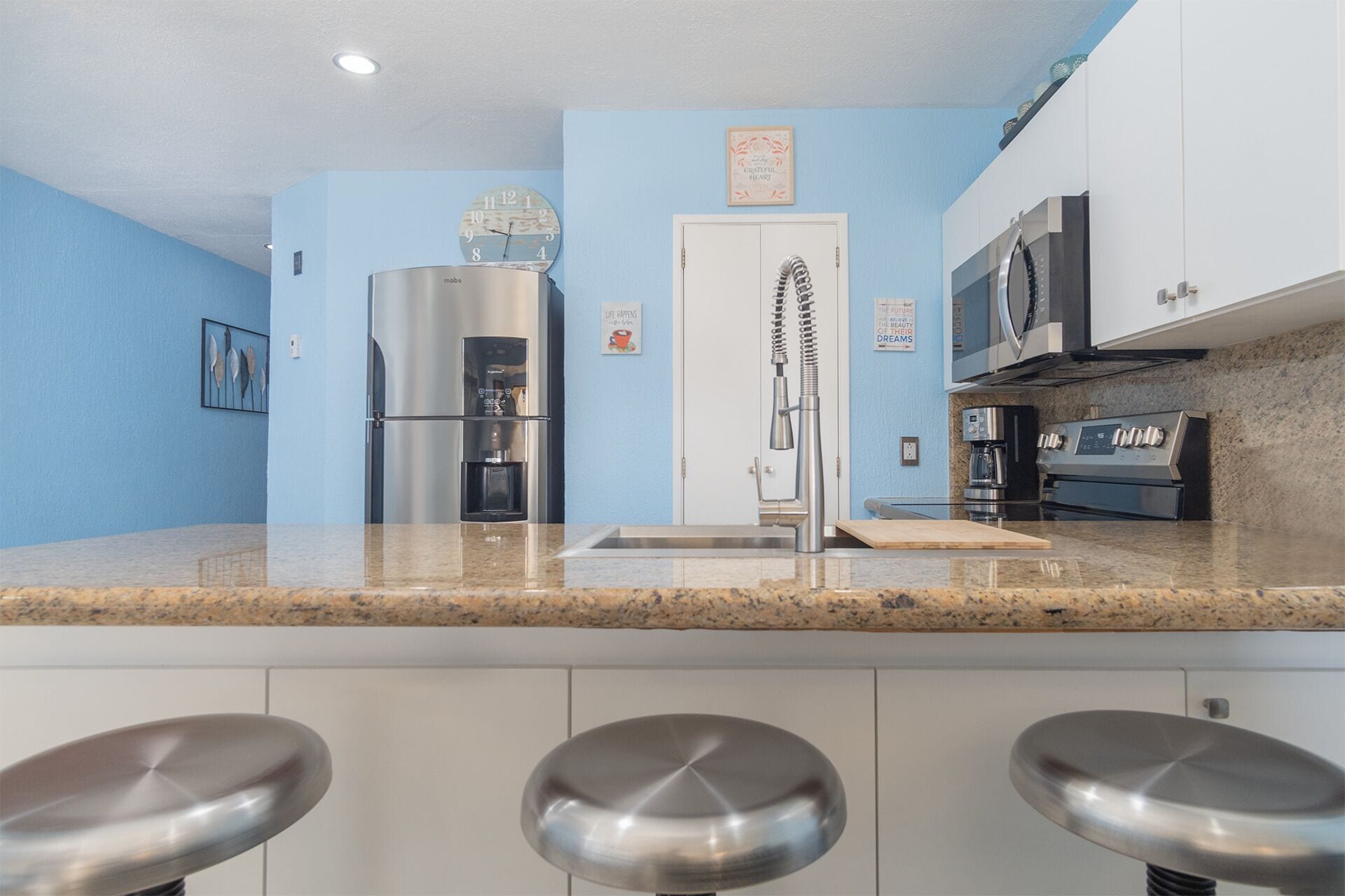 A modern kitchen with a stainless steel refrigerator, a coffee maker, a microwave, and a sink. There are three bar stools in front of the sink.
