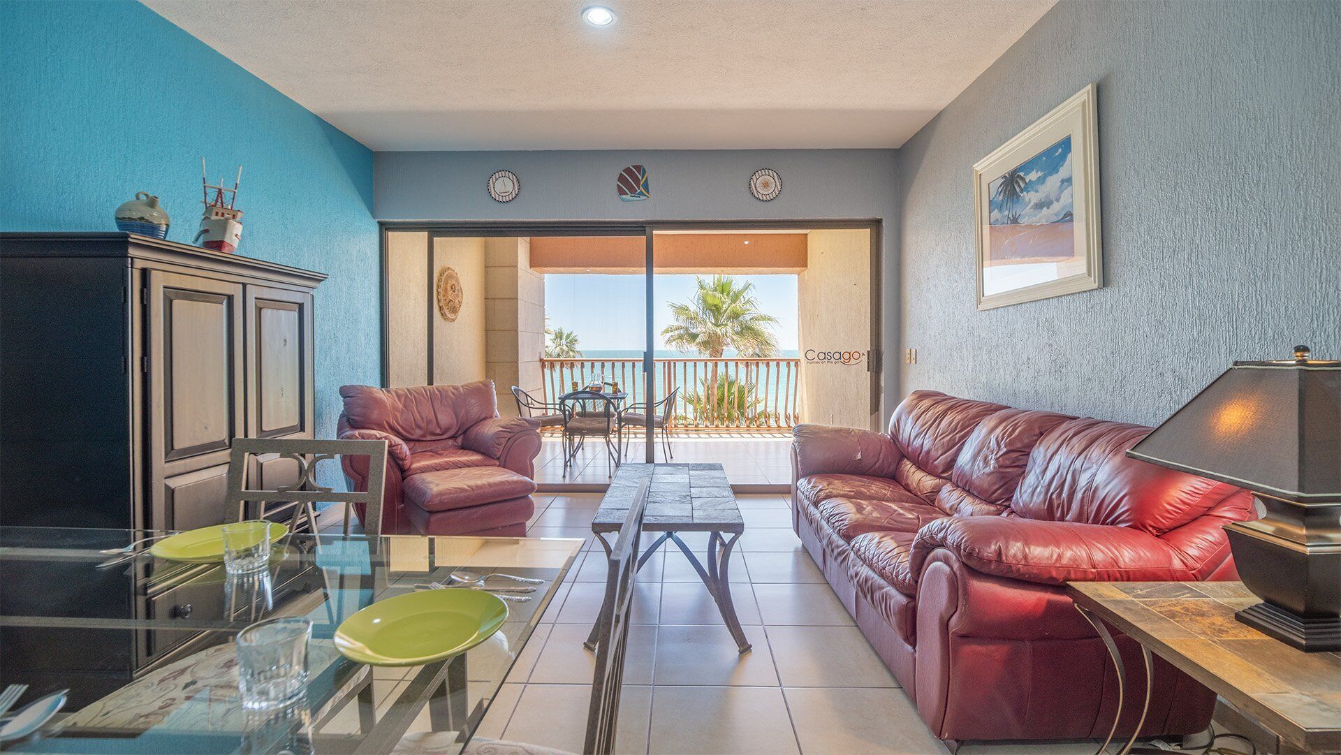 A living room with a glass table, two leather sofas, a cabinet, and a balcony with a view of the sea.