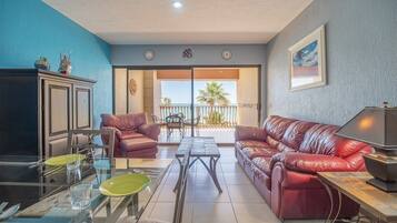 A living room with a glass table, two leather sofas, a cabinet, and a balcony with a view of the sea.