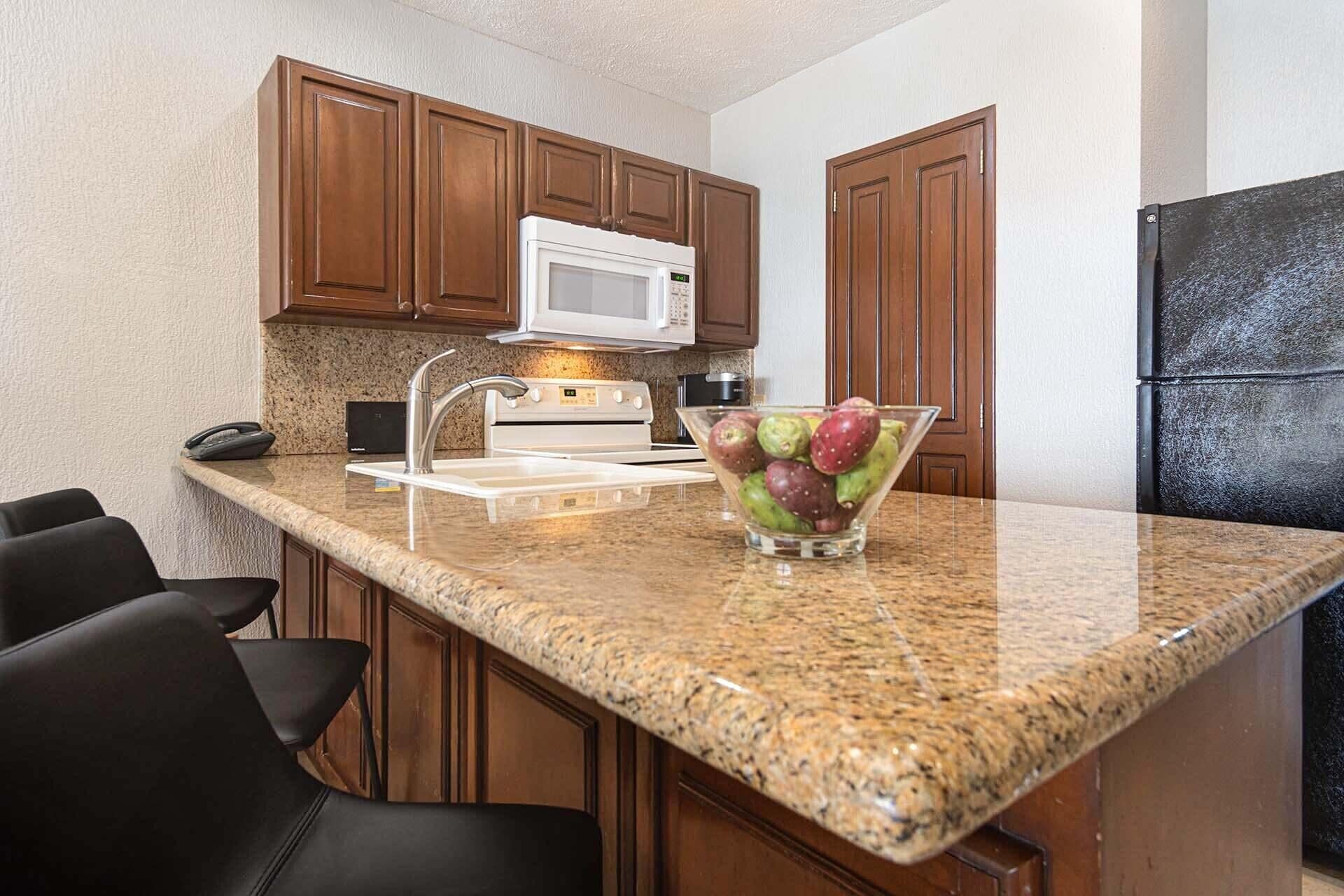 A kitchen with granite countertops, wooden cabinets, a microwave, a refrigerator, and a bowl of fruit.