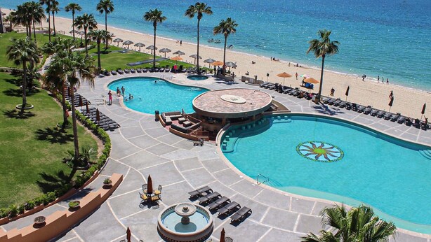 Un resort con dos piscinas amplias, una playa y palmeras.