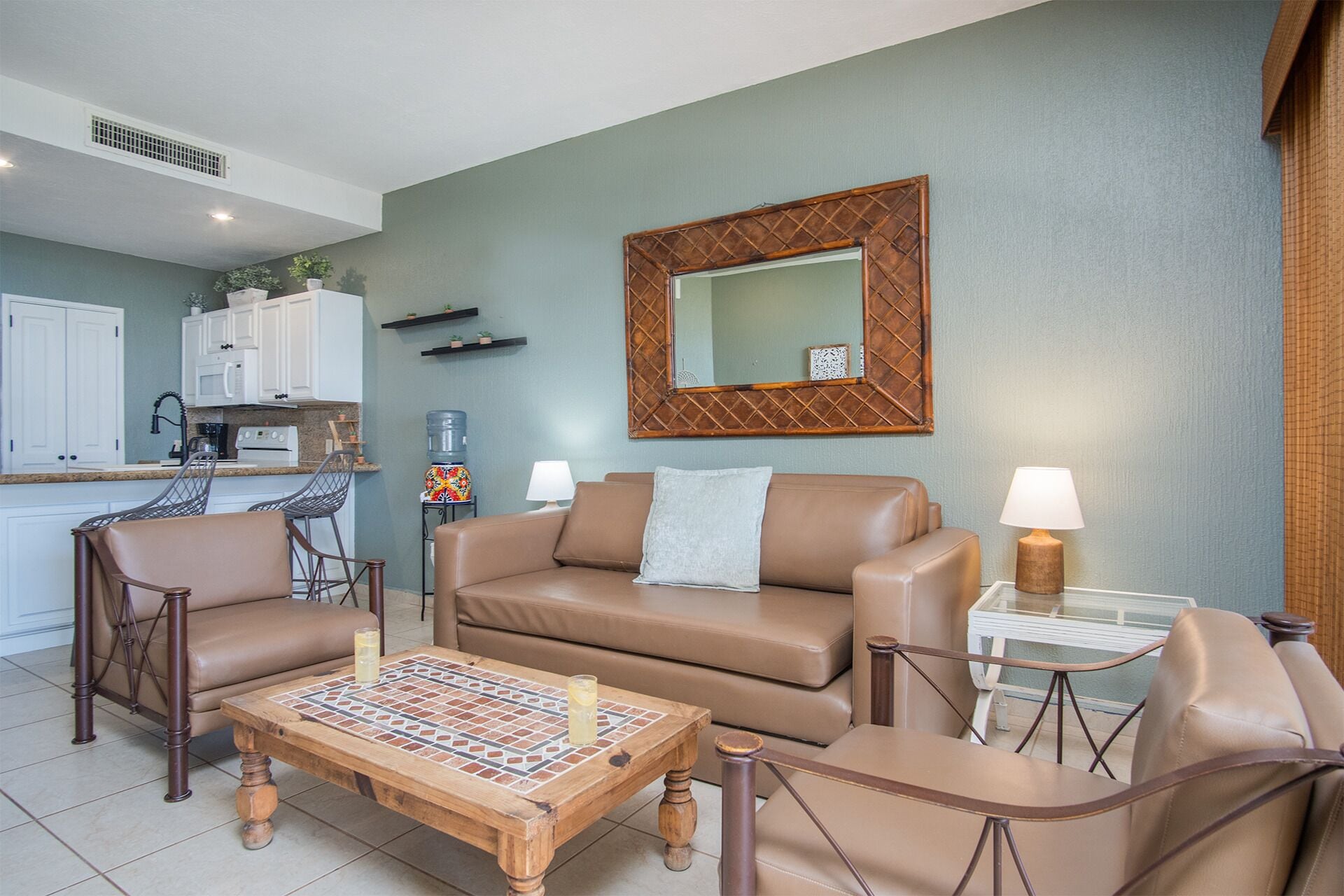 A living room with a brown leather sofa, two armchairs, a coffee table with a mosaic top, a kitchen in the background, and a large mirror on the wall.