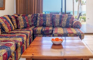 Un salon avec un canapé aux couleurs vives, une table basse en bois sur laquelle se trouve un saladier rempli d’oranges, et une vue sur la mer par une porte vitrée.