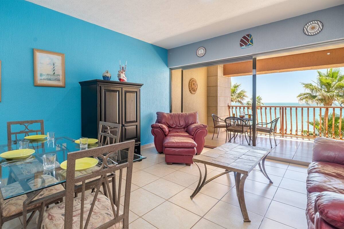 Una habitación con una pared azul, una mesa de comedor con platos amarillos, un sofá rojo y un balcón con vistas al mar.