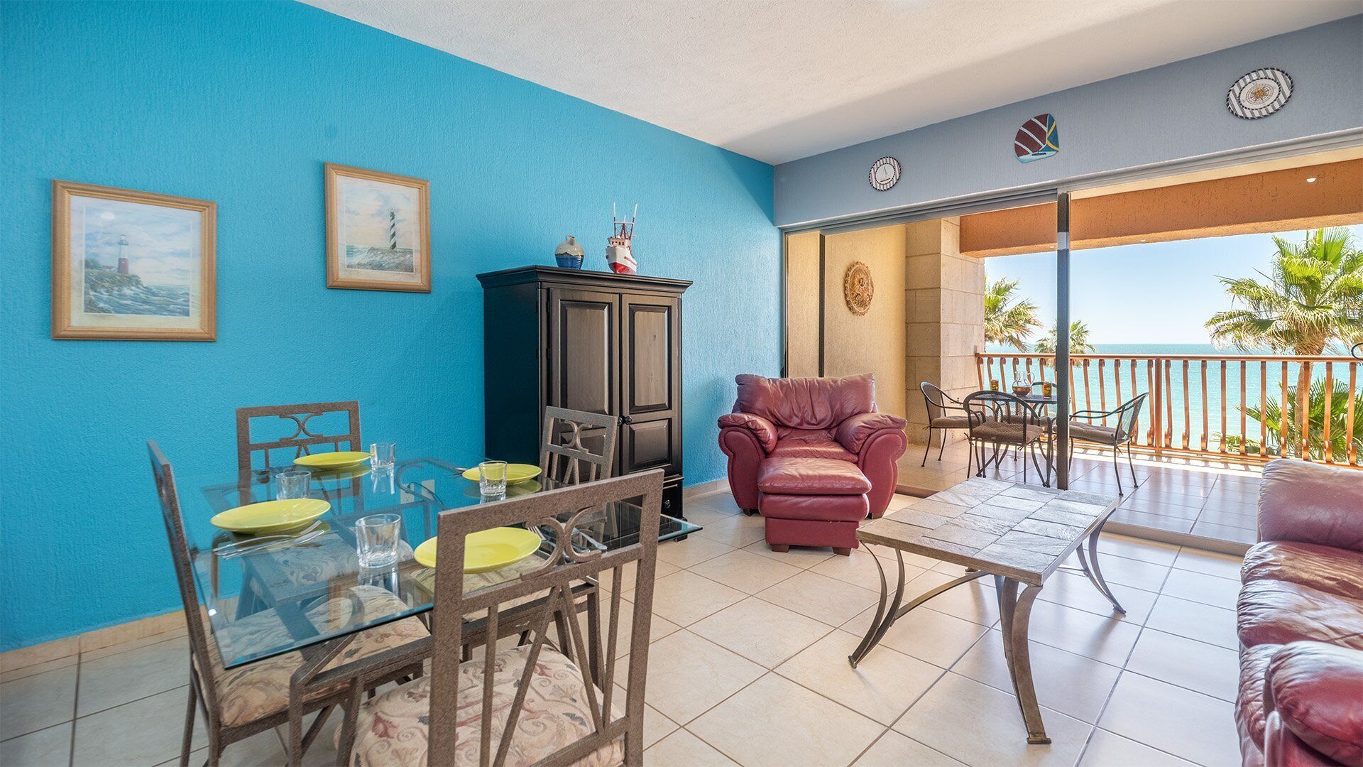 A room with a blue wall, a dining table with yellow plates, a red sofa, and a balcony with a view of the sea.