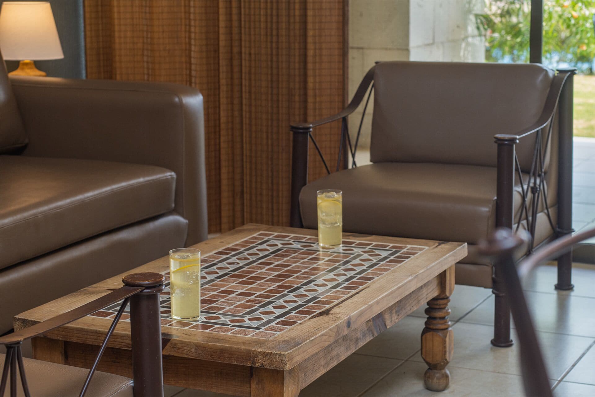 A wooden coffee table with two glasses of a yellow beverage, a lamp, and two brown leather sofas in a room with a tiled floor and a view of greenery outside.