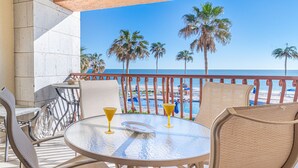 Mesa de vidrio redonda con dos vasos de cóctel amarillos, una silla de mimbre y vista a una playa con palmeras y una piscina.