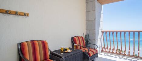 Un balcon avec deux fauteuils en osier, une petite table avec une boisson et des fruits, et une vue sur l’océan.
