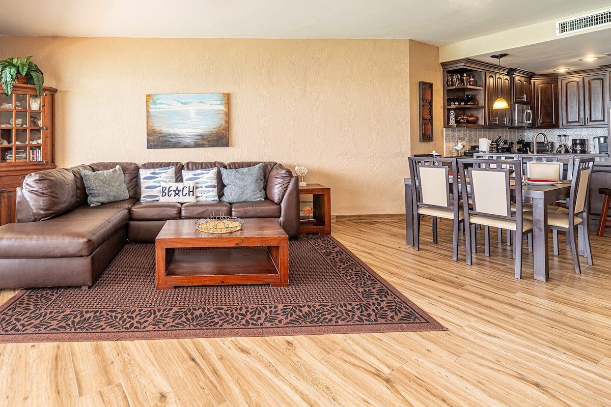 Uma sala de estar com sofá seccional, mesa de centro e tapete. Há um quadro na parede e uma área de cozinha visível ao fundo.
