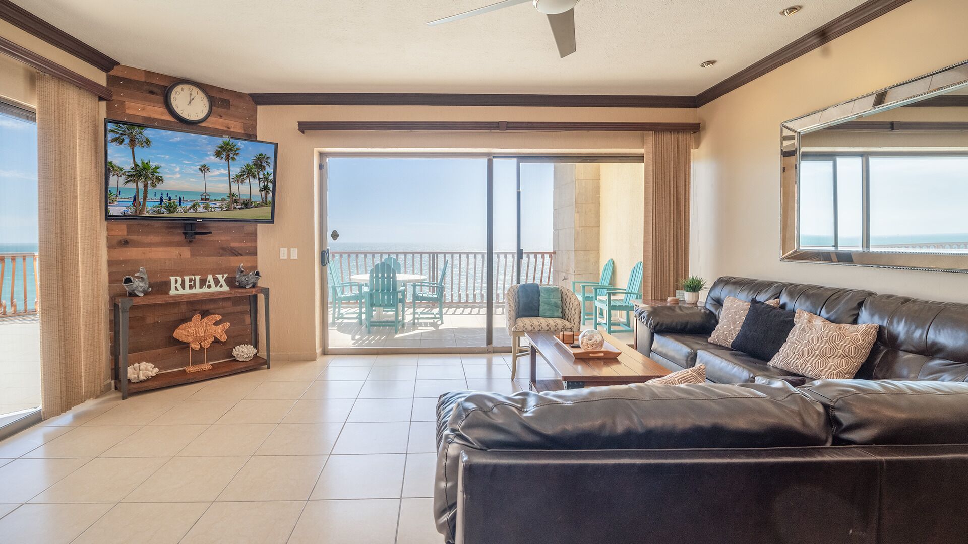 Een ruime woonkamer met een zwarte leren bank, een houten console tafel en een grote televisie aan de muur. Door de glazen deuren is een balkon met uitzicht op de oceaan zichtbaar.