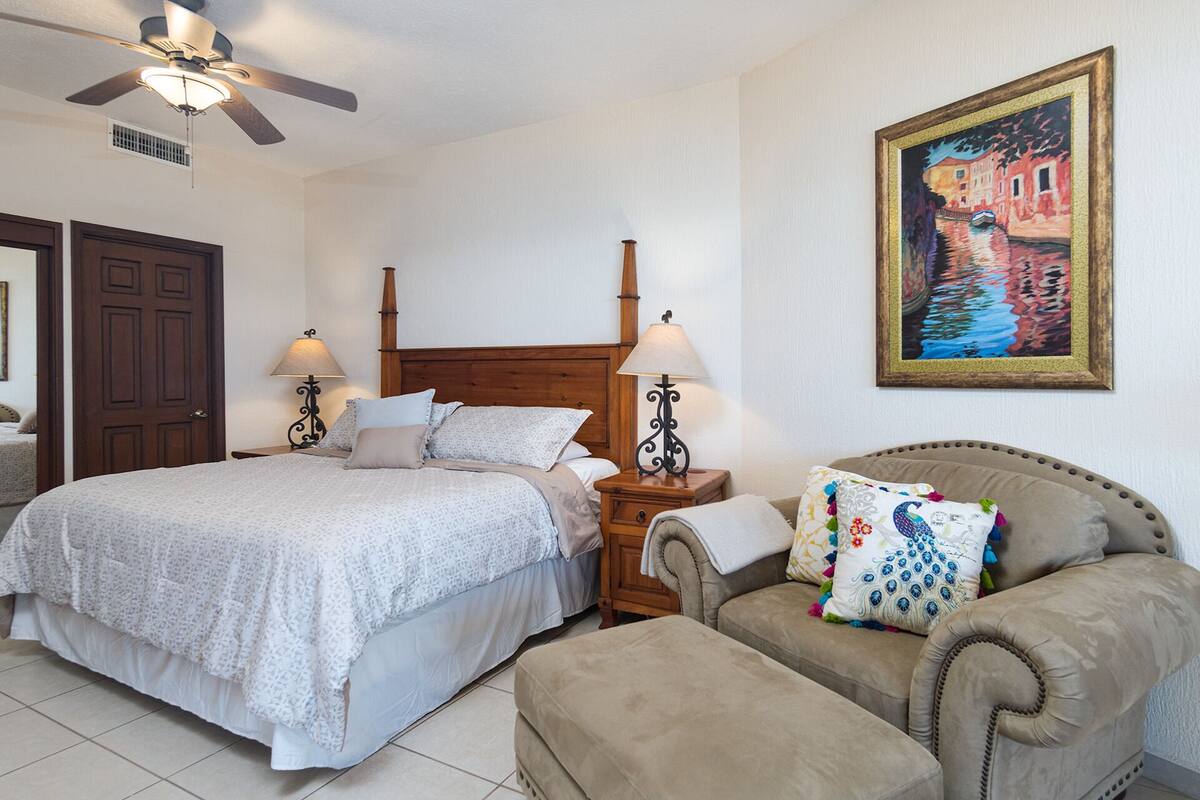 Une chambre à coucher avec un lit double, deux tables de chevet avec des lampes, une tête de lit en bois, une chaise avec un coussin et un tableau au mur.