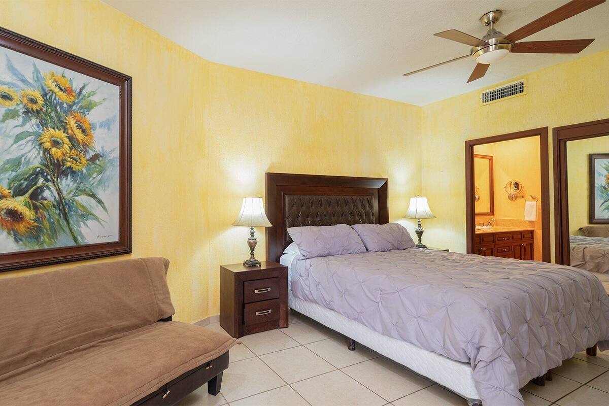 Une chambre d’hôtel avec un grand tableau représentant des tournesols, un lit avec une tête de lit en bois, un canapé marron, deux tables de chevet avec des lampes et un ventilateur de plafond.
