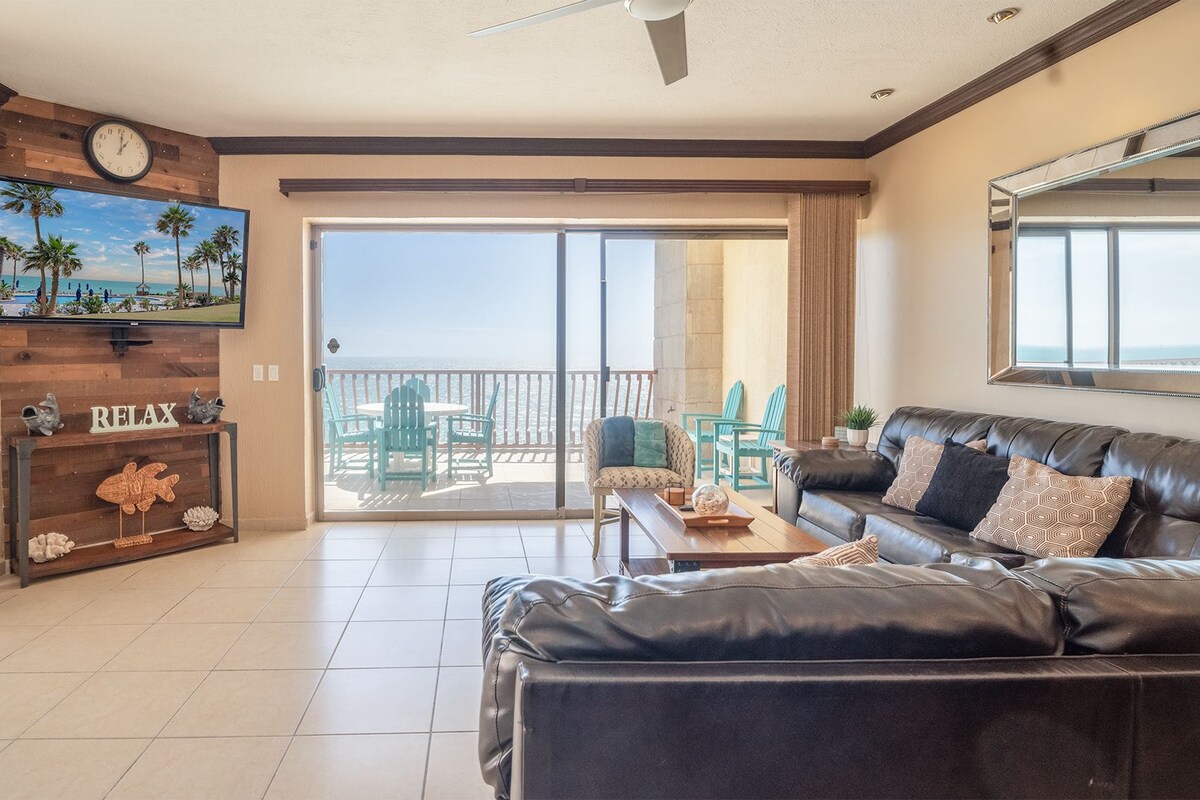 Een ruime woonkamer met een zwarte leren bank, een houten console tafel en een grote televisie aan de muur. Door de glazen deuren is een balkon met uitzicht op de oceaan zichtbaar.