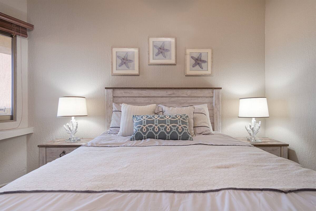 Un lit bien fait, recouvert d’une couvre-lit à motifs, avec deux lampes de chevet et trois étoiles de mer encadrées accrochées au mur.