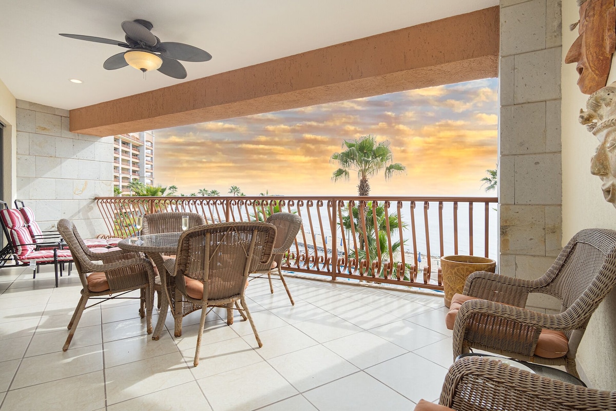 A balcony with wicker furniture, a ceiling fan, and a view of palm trees and a sunset.