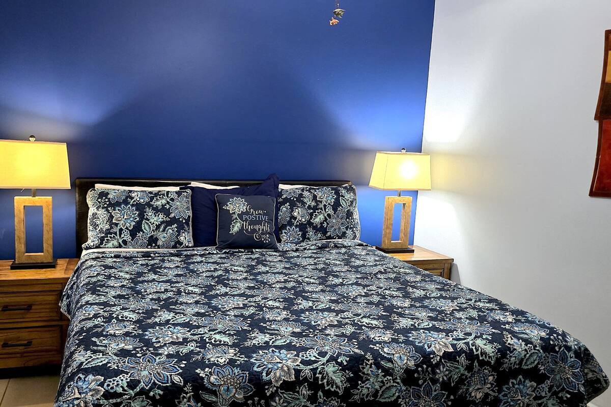 Une chambre avec un mur bleu, un lit recouvert d’une literie à motifs floraux, deux tables de chevet avec des lampes et une commode en bois avec des tiroirs.