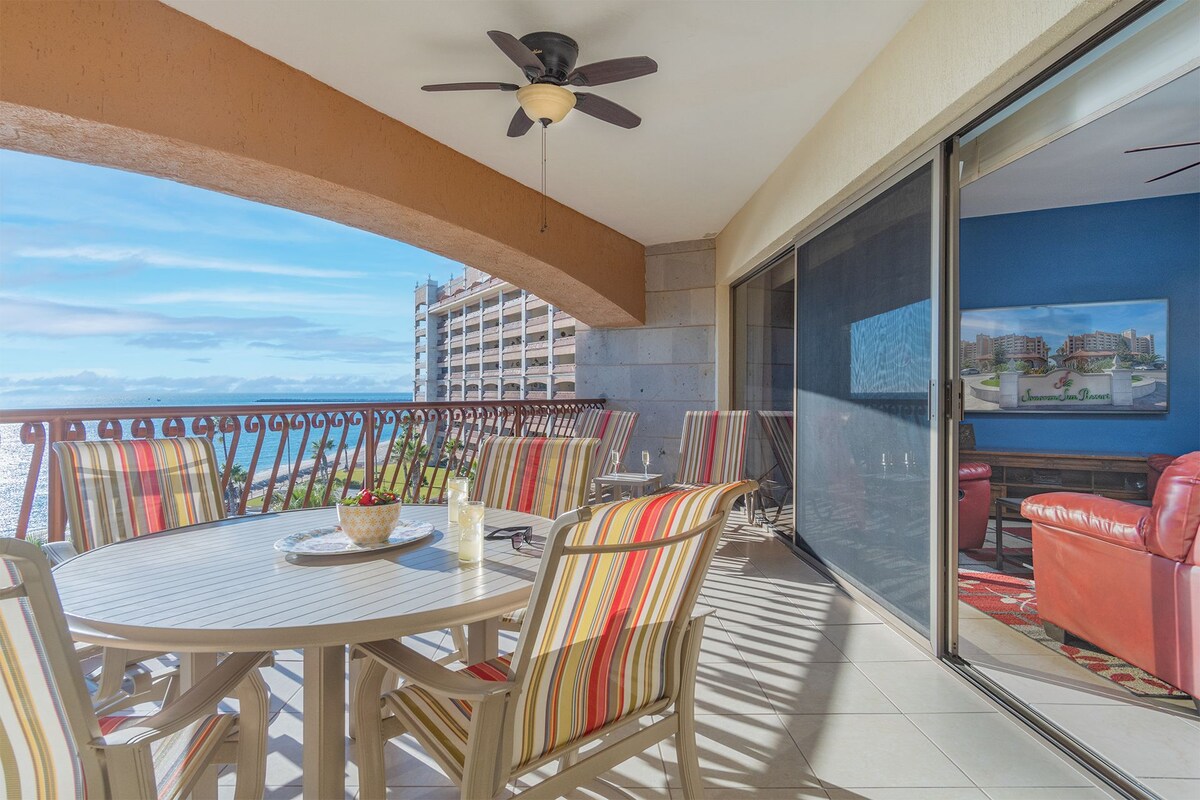 Un balcon avec une table et des chaises, un ventilateur de plafond et une vue sur l’océan et les bâtiments.