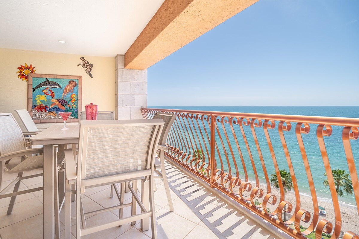 Ein Balkon mit Blick auf das Meer, einem Tisch mit Stühlen und einem farbenfrohen Wandgemälde.
